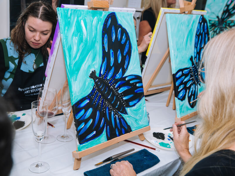 Two women paint blue butterflies at a paint and sip class.