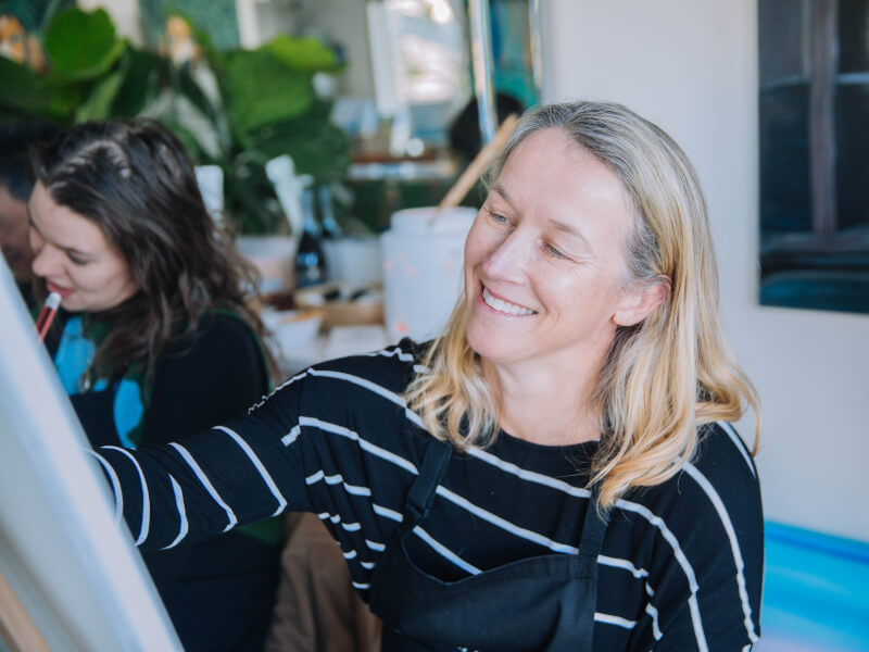Woman smiling whilst painting at a paint and sip class.