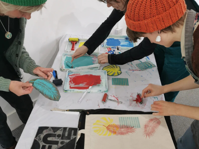 A group of people are standing at a table, applying ink to carved lino blocks to print