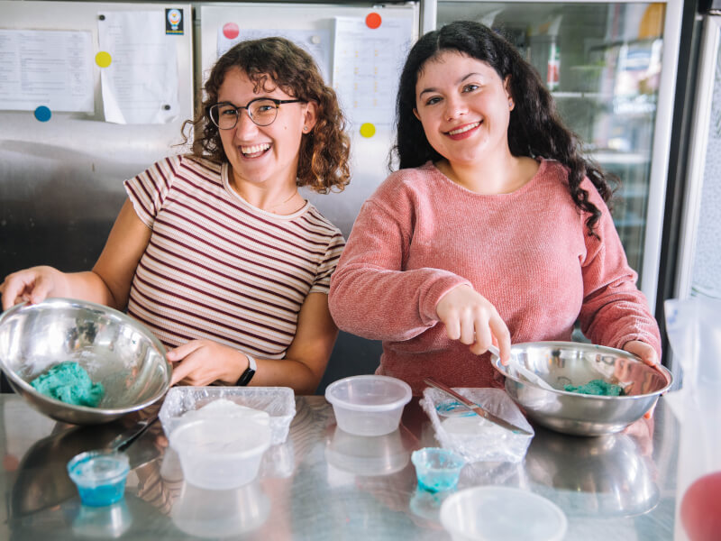 Two women smile widely whilst holding metal bowls containing a blue macaron mixture in a kitchen setting.