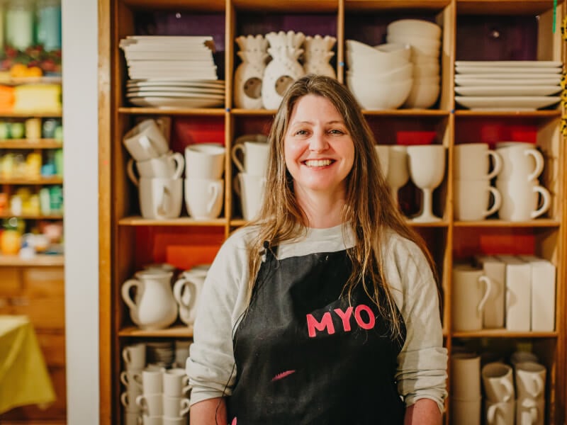A woman wearing an apron smiling in front of shelves of pottery.