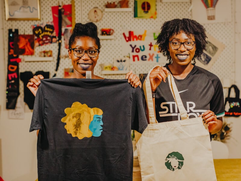 Two women are holding up a tshirt and a tote bag that they've designed using vinyl printing