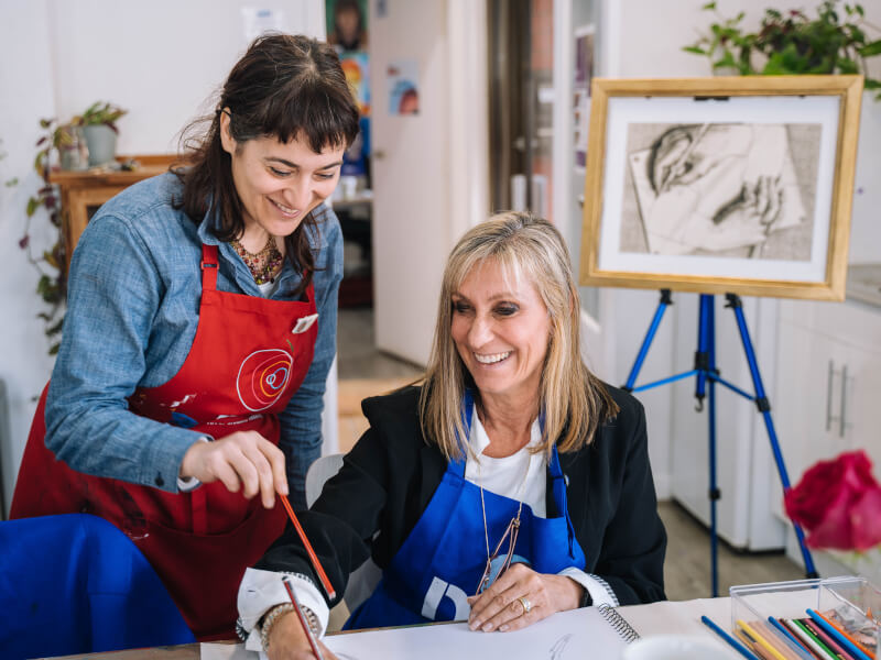 Two women are laughing at a drawing class in Manchester, one is holding a pencil
