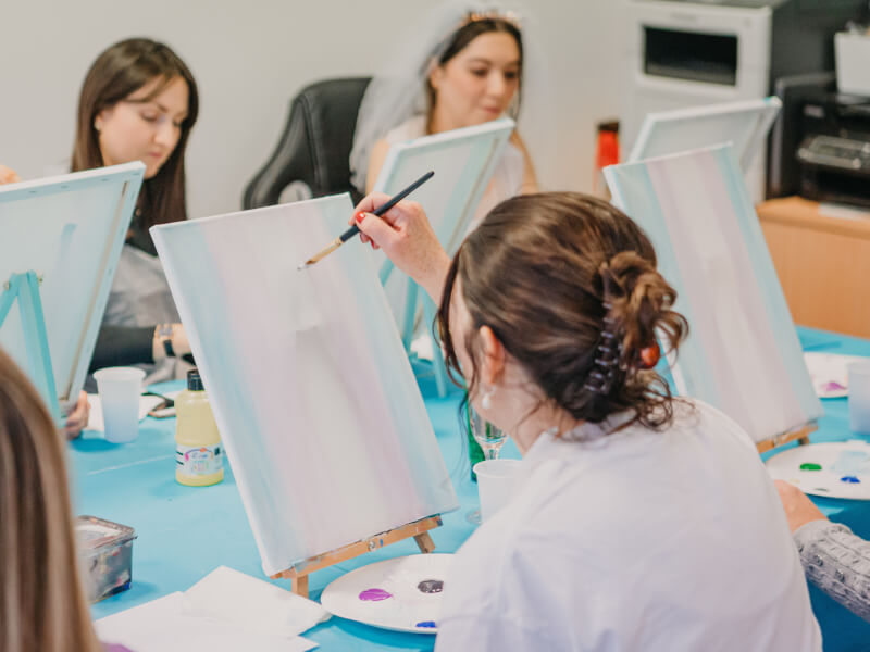 A group of women are sitting at a table in front of easels, painting at a hen party paint and sip class