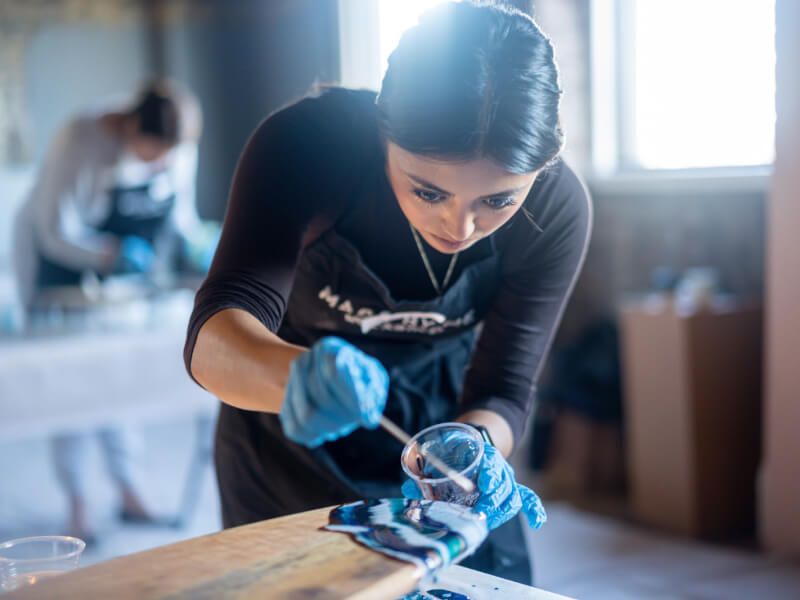 A woman is decorating a chopping board by pouring mixed paint over the end