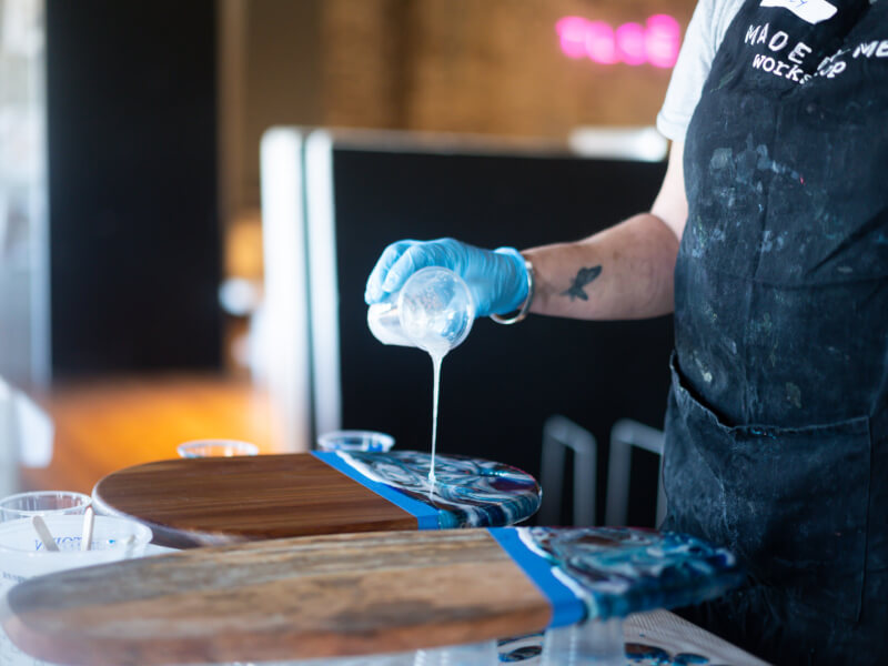 A close up of a person at a paint pouring art class is pouring blue paint on a wooden chopping board