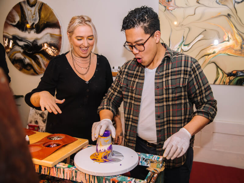 Two people are laughing and creating a fluid artwork using yellow, purple and grey paint