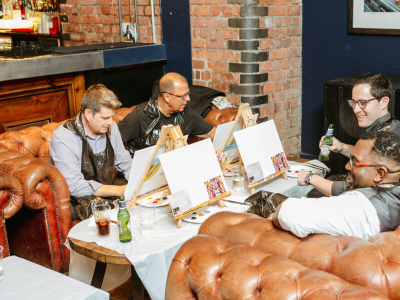 A group of people at an Edinburgh art class are sitting around a table with small table easels, sketching