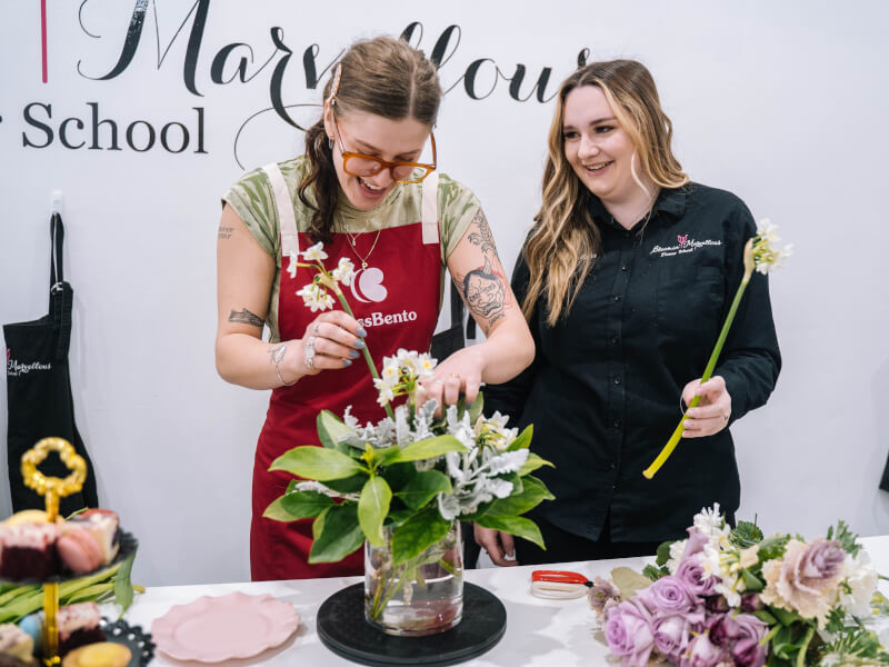 Two people in aprons are making a floral display in a vase from white flowers and green foliage
