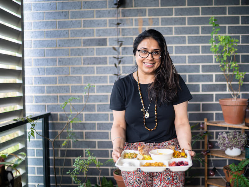 A woman stands on a balcony filled with plants holding a segmented plate filled with various vegan Indian dishes