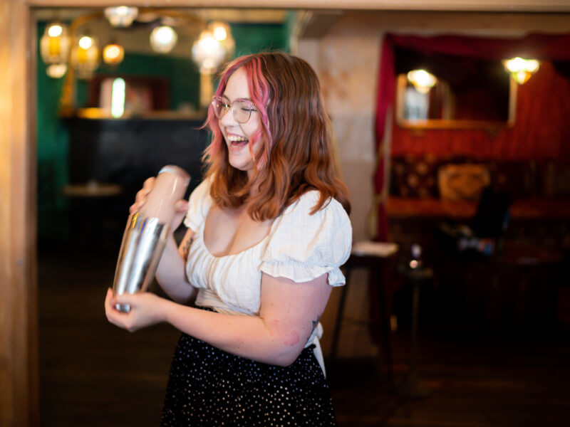 A woman in a white top smiles as she shakes a cocktail in a traditional cocktail shaker