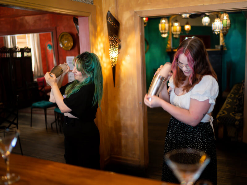 Two women stand side by side behind a bar shaking cocktails