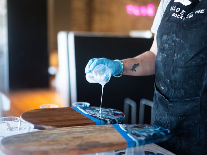 A woman pours acrylic paint onto a wooden board in a fluid art class.