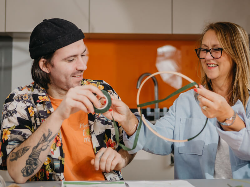 A man and woman are sitting at a table, making a floral hoop decoration from fresh flowers
