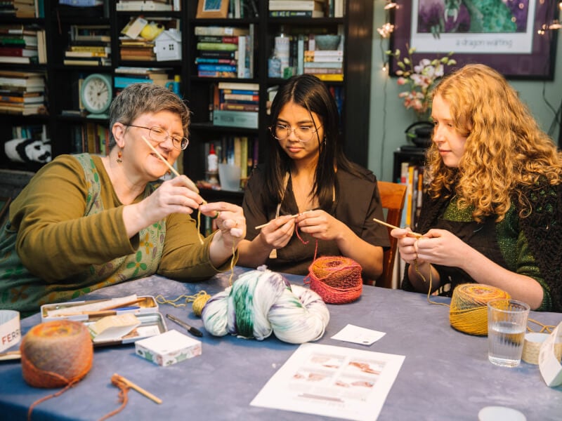 A group of people follow a teacher as they guide them through a crochet class.