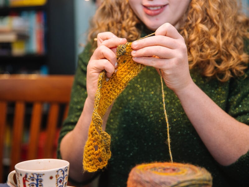A person knitting with green and orange yarn with a cup of tea to the left 