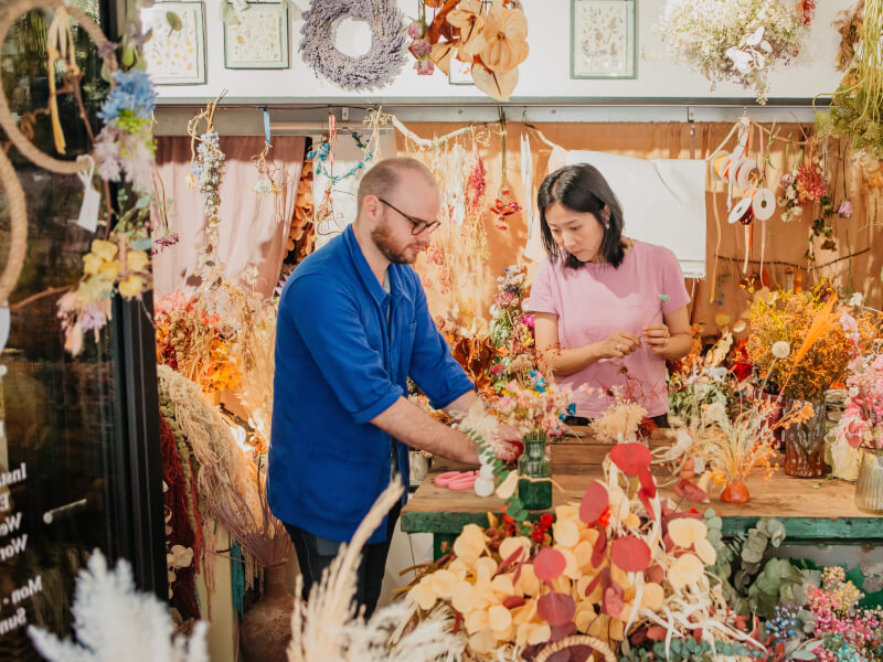 A man and woman are making floral arrangements at a wooden table in a room filled with dried flowers