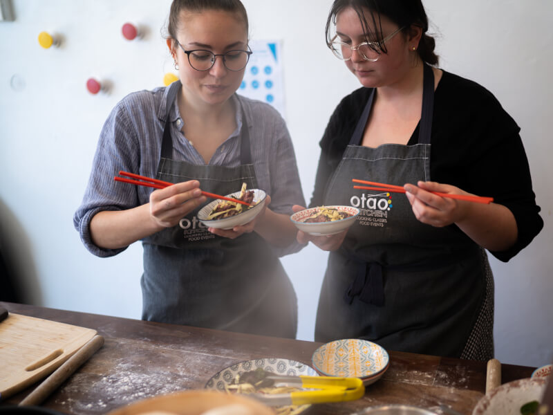 Two women eating out of small bowls with chopsticks at a cooking class.