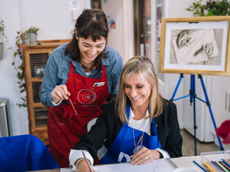 A woman in a red apron is teaching a second woman how to paint