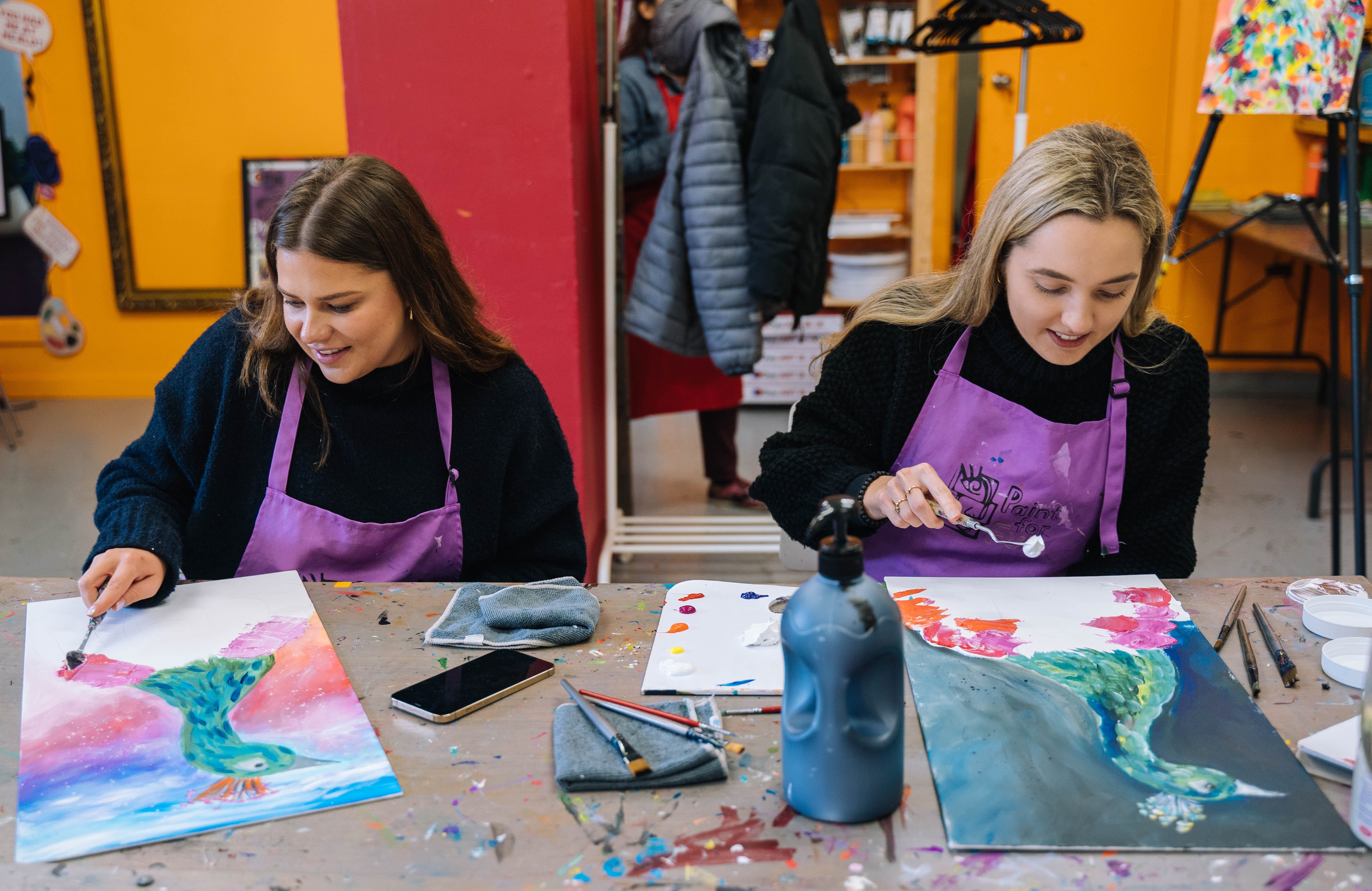 Two women in purple aprons sit at a paint and sip class