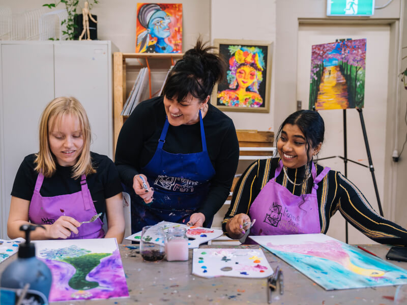 A woman is standing between two other women who are sitting down, teaching them to paint using a pallette knife