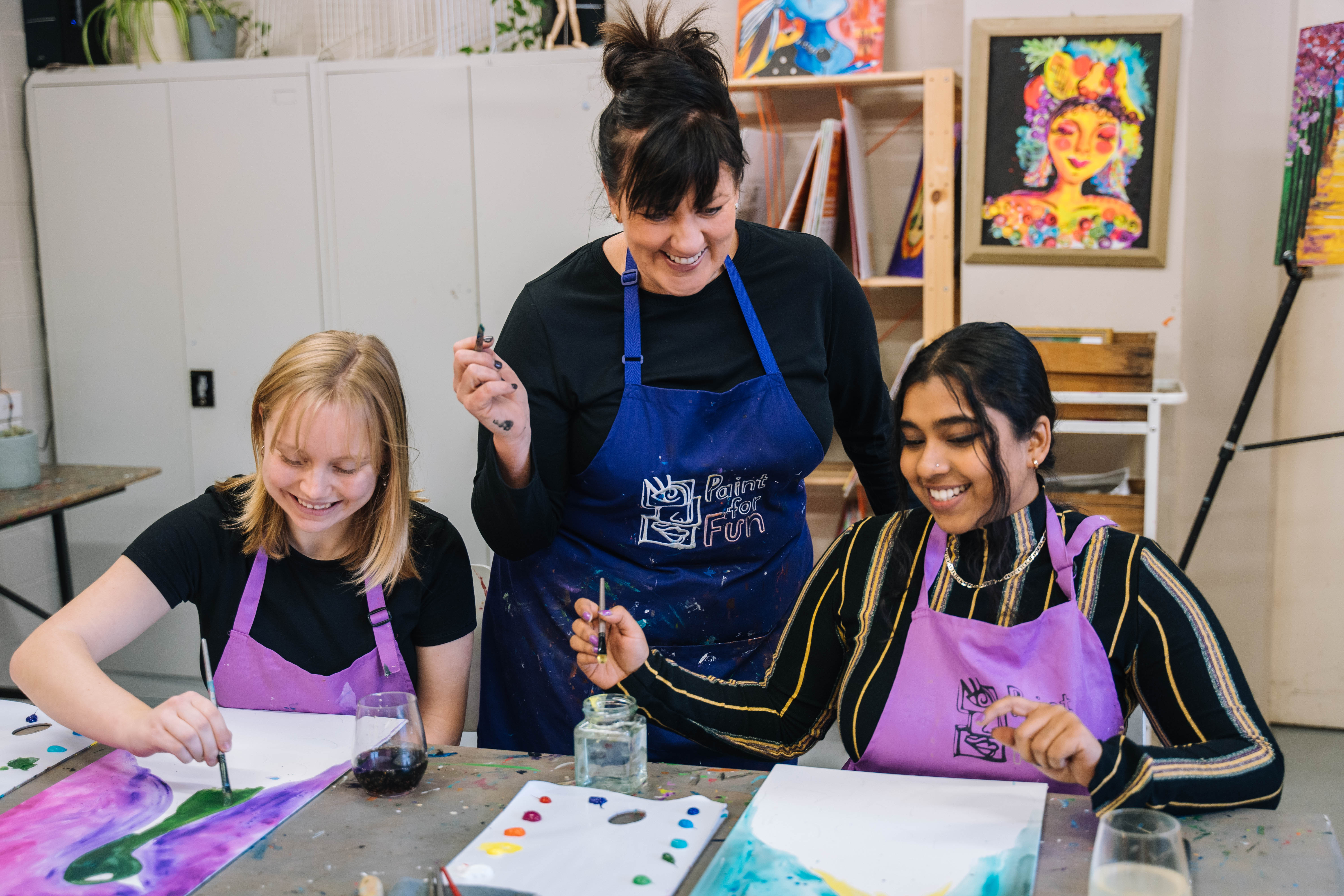 Two smiling women sit side by side painting, while an art teacher supervises them