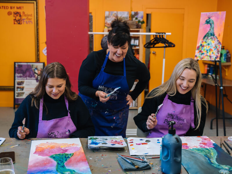 Two women smiling and wearing aprons at a paint and sip class.
