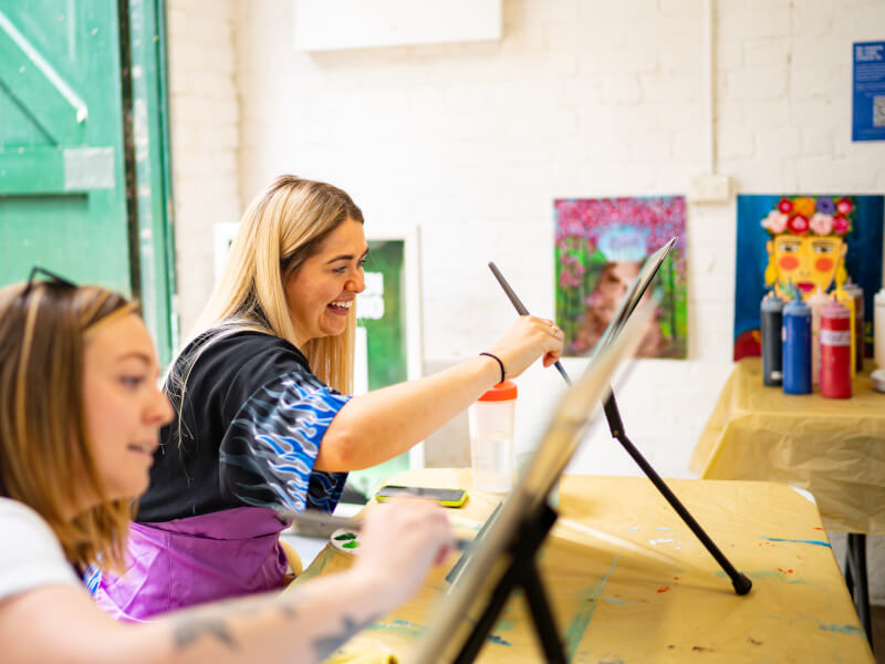 A woman holding a brush sat at a paint canvas.