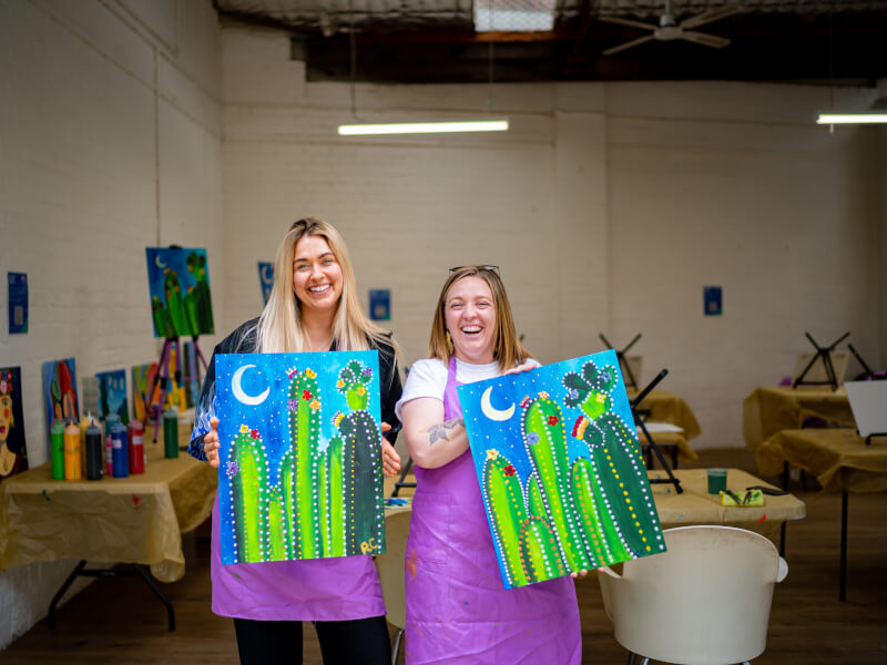 Two women hold up paintings of cactus' that they made in a painting class.
