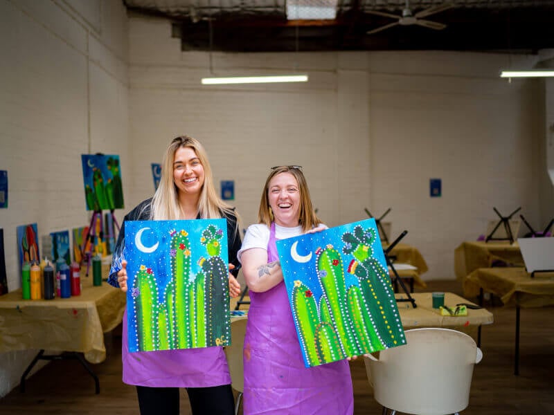 Two women are holding canvases with pictures they've painted on them 
