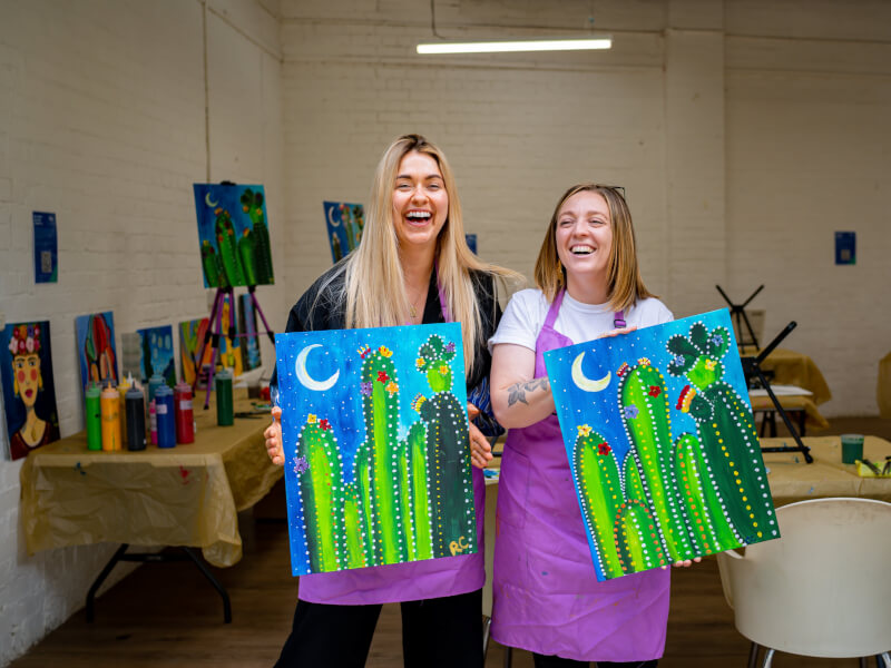Two smiling women holding up artwork at a painting class.