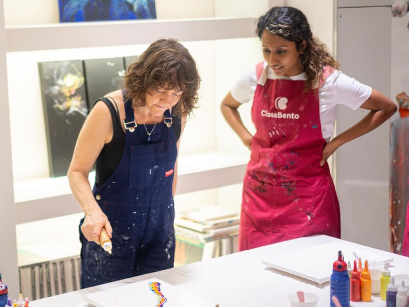 Teacher in paint-splattered overalls demonstrating acrylic pouring by splattering yellow paint on a canvas