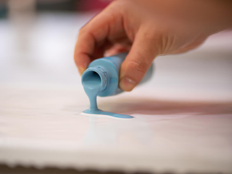 A hand pours a small blue bottle of flowing paint onto a white surface