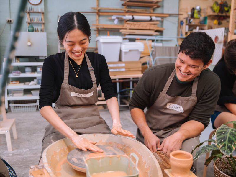 A man a woman smile as they try wheel throwing in a pottery studio.