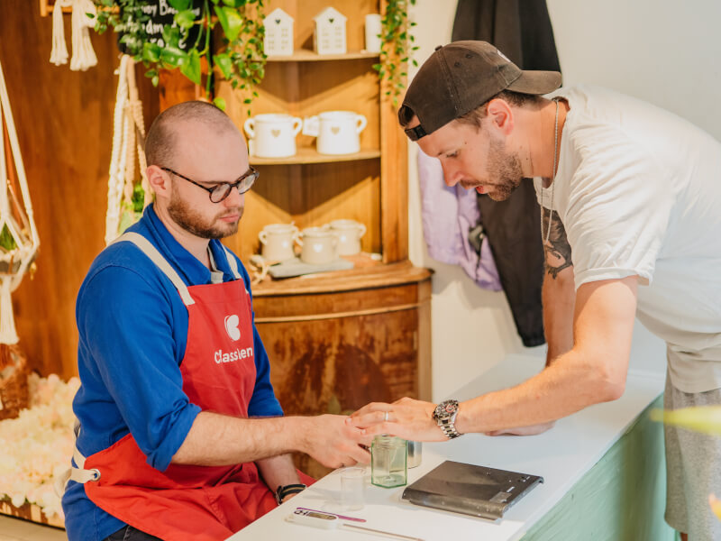 A man sits at a counter as a candle making teacher shows him some skills