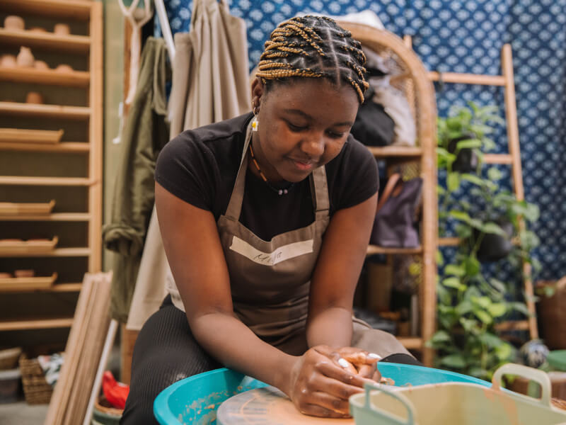 A woman with braided hair a black tshirt and brown apron smiles down at a pottery wheel with both hands clasped around a ball of clay