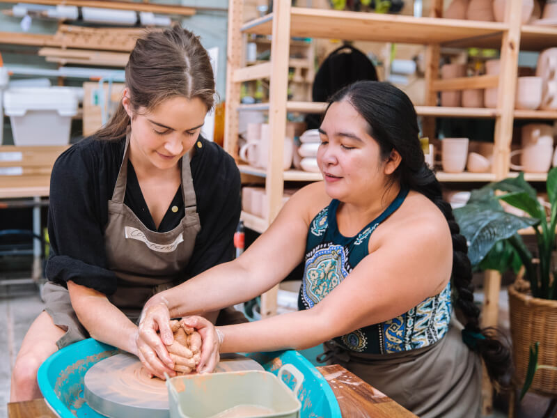 Two women work on a potters wheel, one clasping her hands around the others as she shows her how to mould the clay