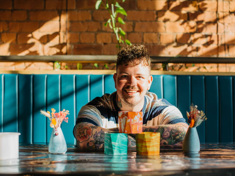 A man is sat behind a wooden table with handmade colourful geometric clay pots stacked in front of him