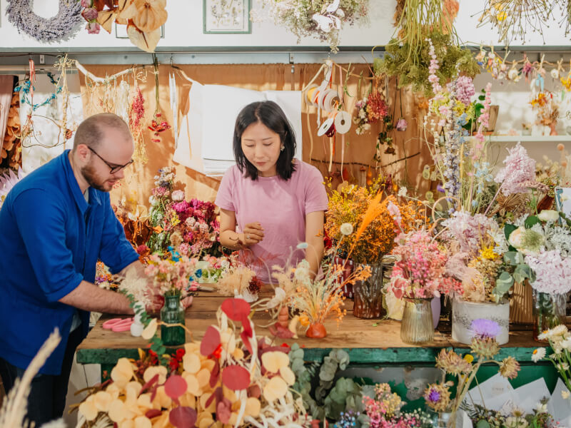 A man and woman standing at a workbench surrounded by bright dried and fresh flowers 