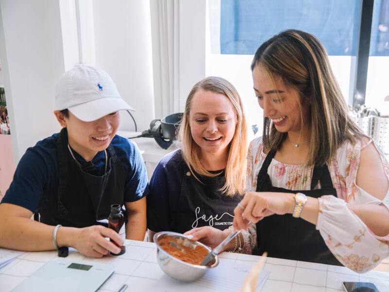 Three women are sitting at a table and mixing the ingredients for a natural face mask in a bowl