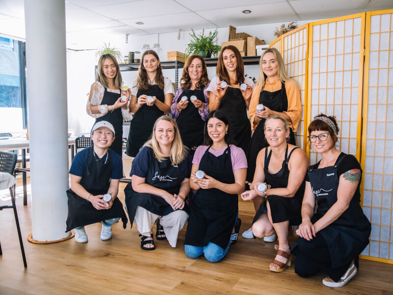 A group of women wearing aprons smiling whilst holding DIY candles.