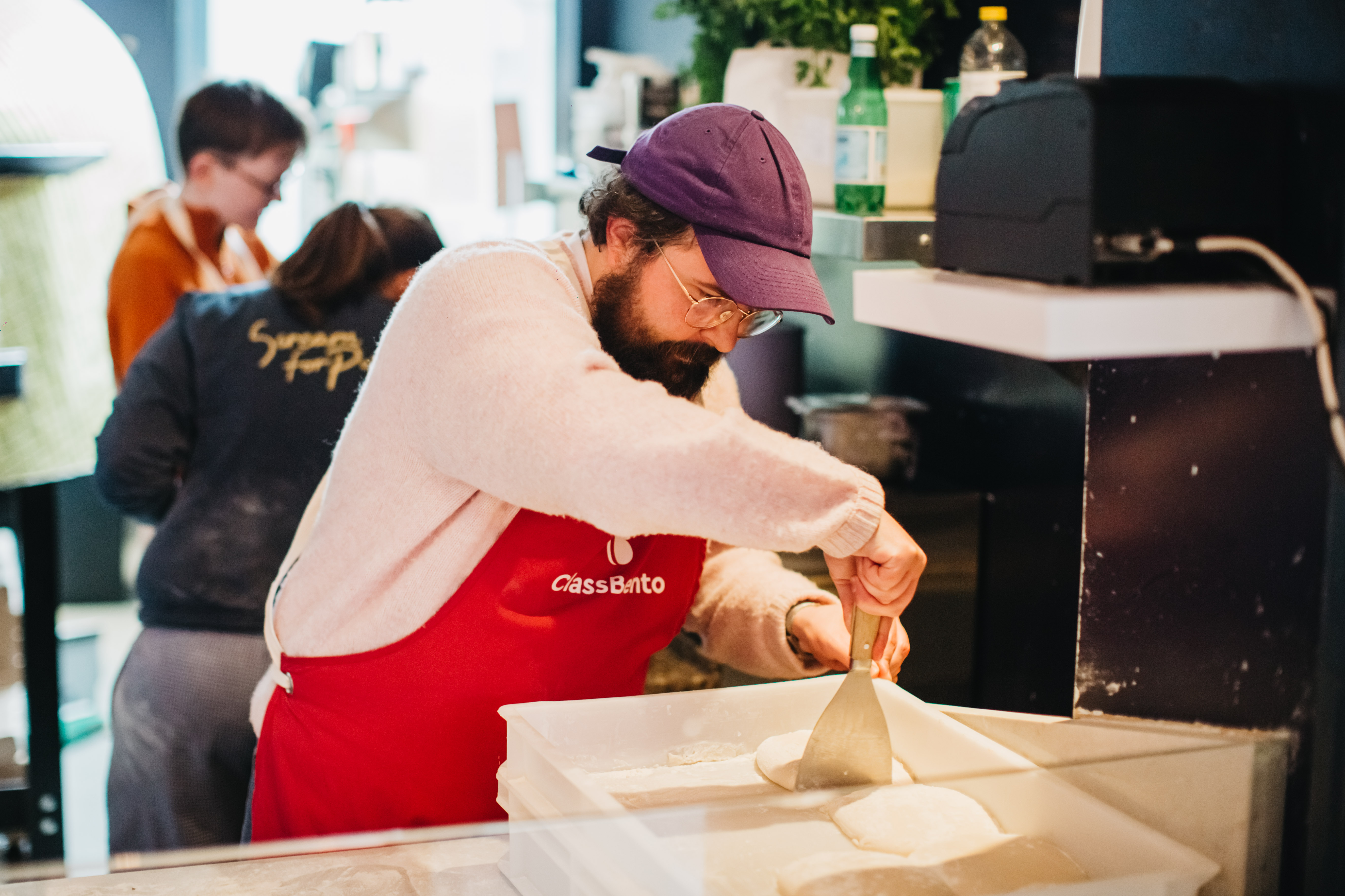 A person in a purple cap taking pizza dough from a tray with a dough scraper
