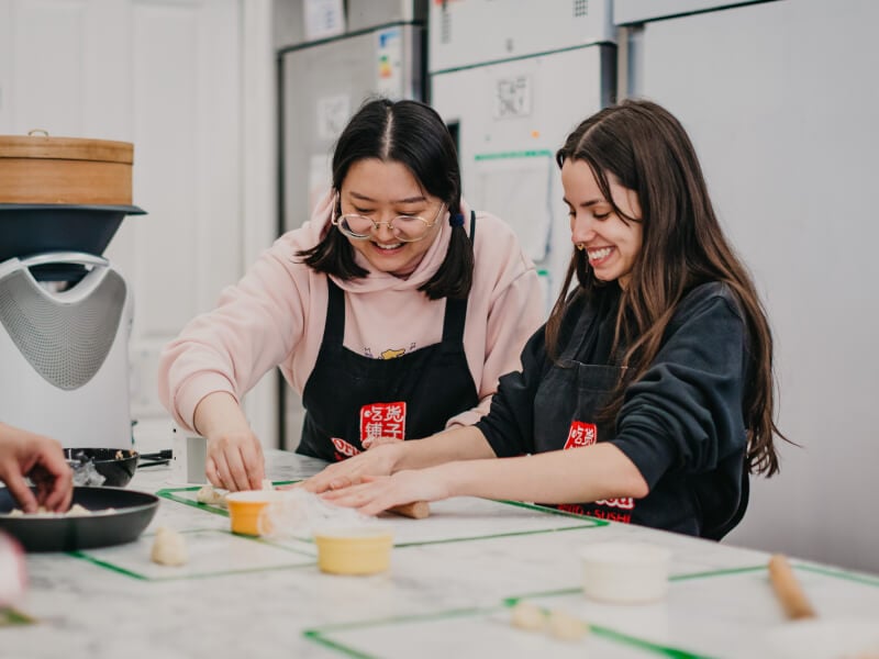 Two smiling women rolling dough for homemade dumplings.