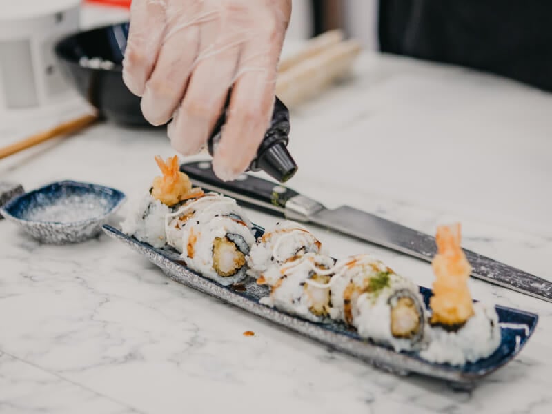 Close up of a person adding seasoning to a row of sushi rolls