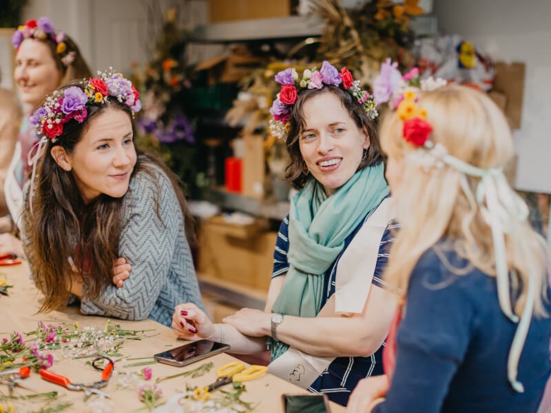 A group of women sit at a wooden table wearing flower crowns