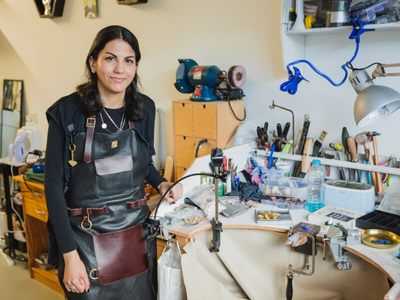 A woman wearing a leather apron stands in a jewellery making studio surrounded by tools and equipment