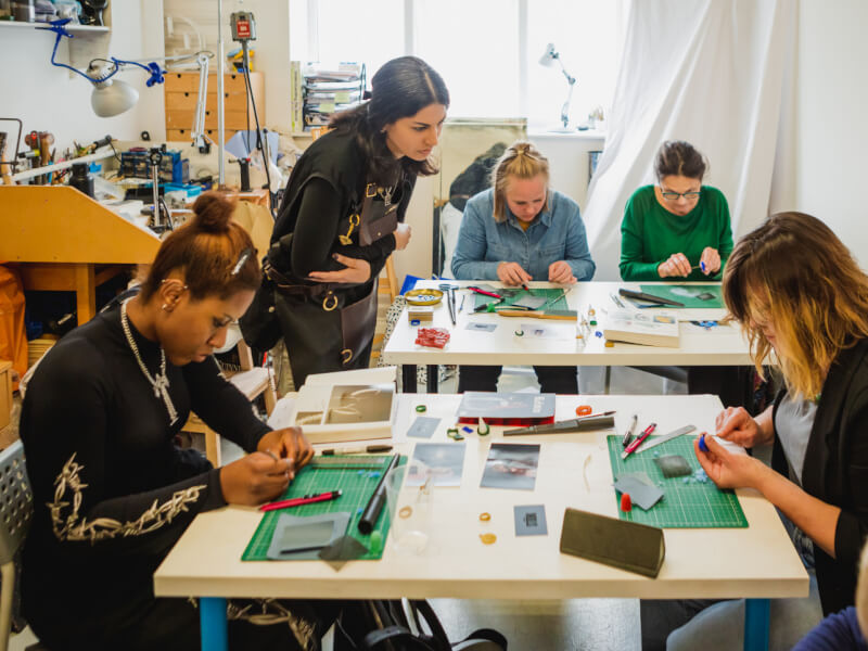 A woman stands above another woman sat at a table, teaching her how to make jewellery