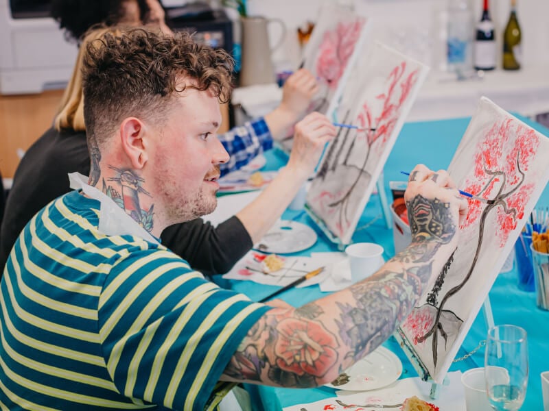 A man with curly brown hair wearing a green and white striped tshirt paints a cherry blossom tree on an almost finished canvas 