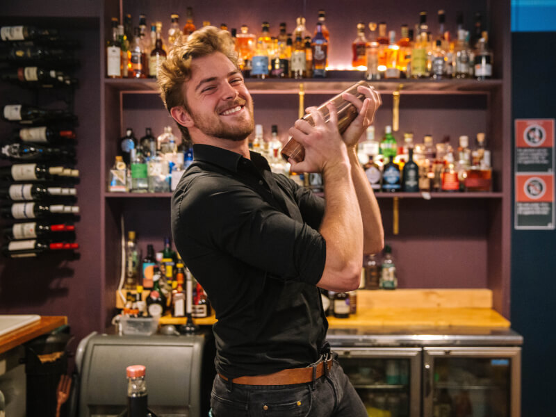 A man shaking a cocktail and smiling behind a bar.