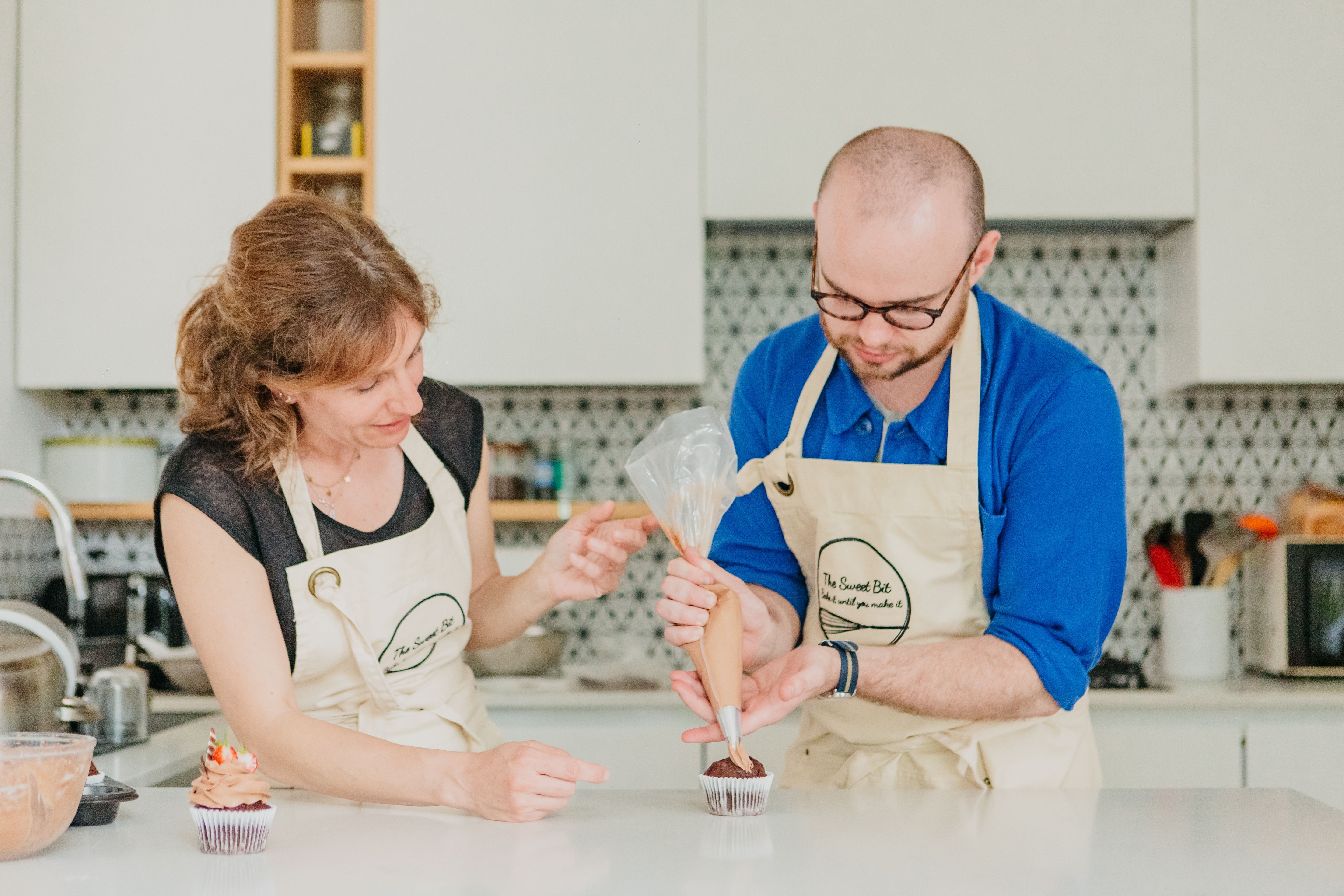 Two people are decorating a cupcake with buttercream frosting and a piping bag